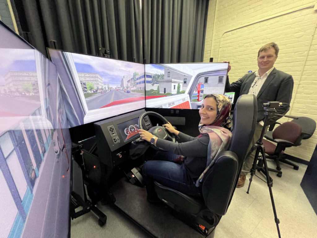 This image shows the quarter-cab truck driving simulator . The image shows the entire setup which is composed of 3 monitors, steering wheel and pedals. Also, in the image Prof. Roorda and doctoral student Alia Galal appear